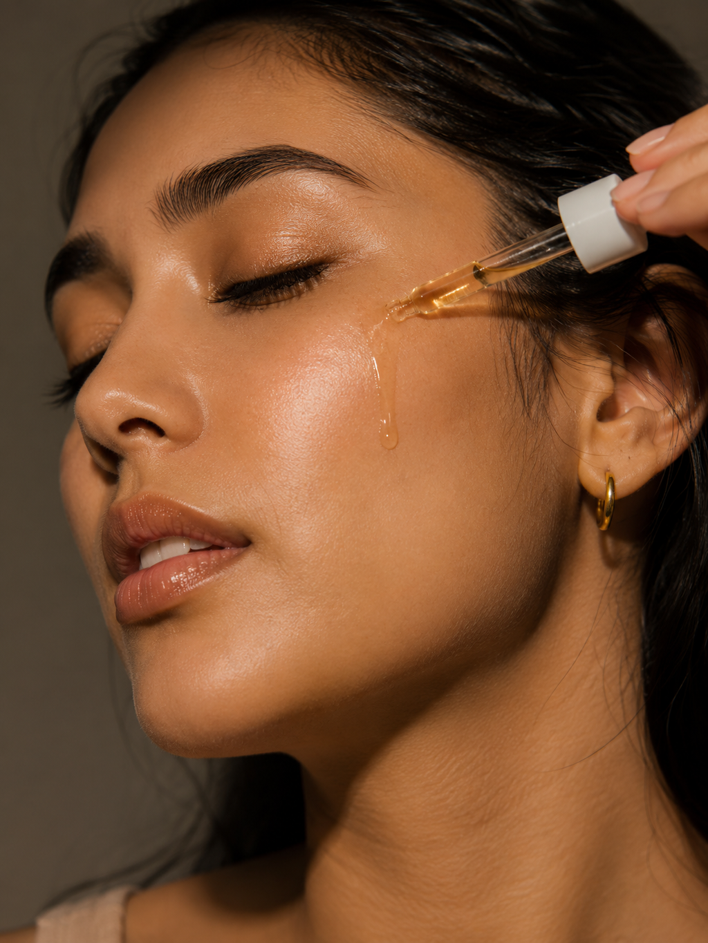 Close-up of a woman applying a golden facial serum with a dropper, with a single drop of liquid dripping from the dropper onto her cheek.