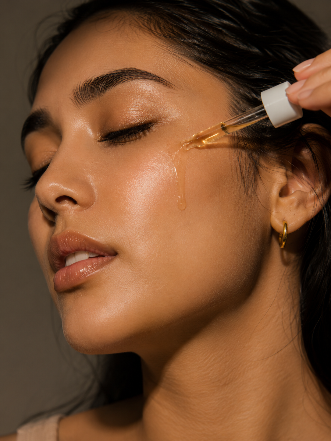 Close-up of a woman applying a golden facial serum with a dropper, with a single drop of liquid dripping from the dropper onto her cheek.